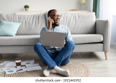 Always In Touch. Black Freelancer Man Talking On Phone Using Laptop Working Remotely From Home, Sitting On Floor In Living Room. Freelance Career, Distant Work And Mobile Communication