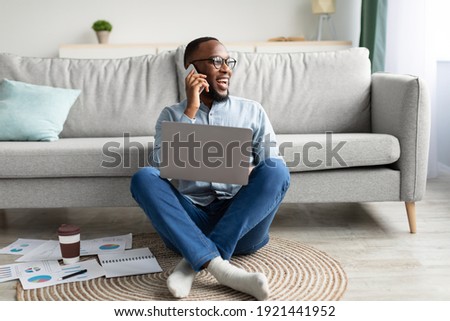 Always In Touch. Black Freelancer Man Talking On Phone Using Laptop Working Remotely From Home, Sitting On Floor In Living Room. Freelance Career, Distant Work And Mobile Communication