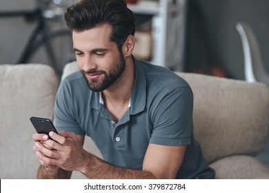 Always in touch. Cheerful handsome young man using his smart phone while sitting on the couch at home
