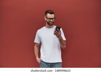 Always in touch. Handsome young man in casual clothes holding mobile phone and smiling while standing against red wall outdoors - Powered by Shutterstock