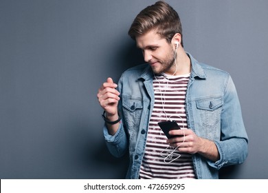 Always In Touch. Handsome Young Man Wearing Headphones And Holding Mobile Phone While Standing Against Grey Wall