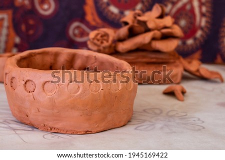 An amateur pinch pot with round circles decorating its rim against a mandala tapestry on parchment paper. Pile of unused terracotta clay in the back, closeup shot in artist workshop.
