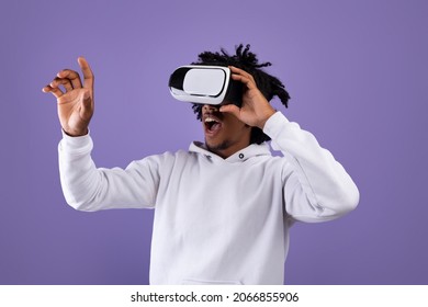 Amazed African American teen guy in VR headset using virtual reality, touching imaginary screen on violet background. Funky black adolescent exploring cyberspace, experiencing modern technologies