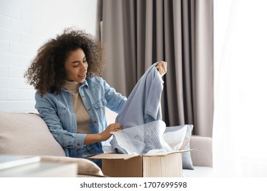 Amazed afro american young woman customer unboxing fashion delivery purchase excited about present in post shipment parcel. Excited african lady shopper opening cardboard box sit on sofa at home.