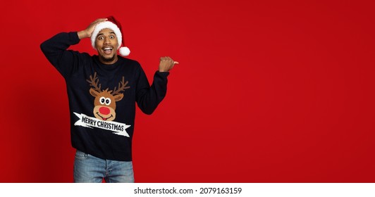 Amazed Black Guy In Red Santa Hat And Blue Sweater With Deer Pointing At Copy Space For Advertisement Over Red Studio Background, Showing Exciting Christmas Deal, Panorama, Web-banner