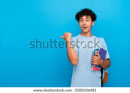 Amazed boy guy high school male pupil student teenager with bag holding books copybooks pointing finger empty copy space for advertisement isolated on blue background. Education back to school concept