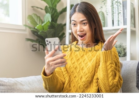 Amazed, brunette asian young woman, girl happy sit on couch hold, using mobile app, read good news online feel excited getting offer, great positive surprise at home,  joy celebrate success on phone.