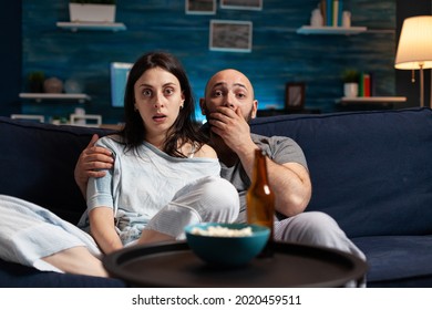 Amazed Confused Couple Watching Movie On TV At Night And Eating Popcorn, Drinking Beer Having Facial Expression. Focused Surprised Concentrated Astonished Wife Sitting Home On Confortable Couch