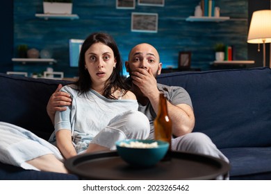 Amazed Confused Couple Watching Movie On TV At Night And Eating Popcorn, Drinking Beer Having Facial Expression. Focused Surprised Concentrated Astonished Wife Sitting Home On Confortable Couch