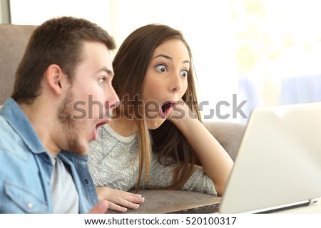 Amazed couple with open mouth on line with a laptop lying on a couch in the living room in a house interior