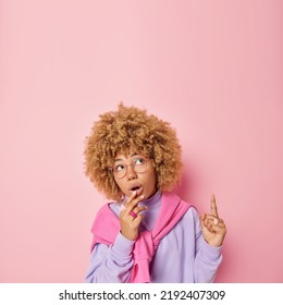 Amazed Curly Haired Woman Points Index Finger Above Shows Awesome Advertisement Stands Fascinated Dressed In Casual Clothes Concentrated Overhead Poses Against Pink Background. Omg Look There