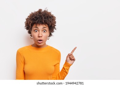 Amazed Curly Haired Young Woman With Shocked Eexpression Has Stunned Face Expression Wears Orange Jumper Points Aside On Blank Space Isolated Over White Background. Copy Space For Advertisement