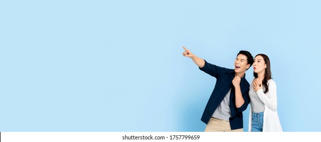 Amazed excited Asian couple tourists pointing hand to empty space on isolated light blue banner background