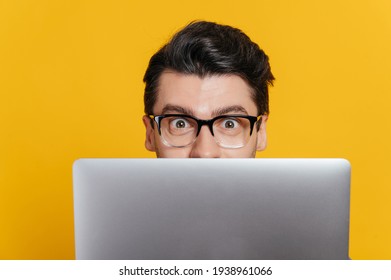 Amazed excited caucasian young adult guy with glasses peeking out from behind laptop, looking surprised at camera while standing against isolated orange background, close-up