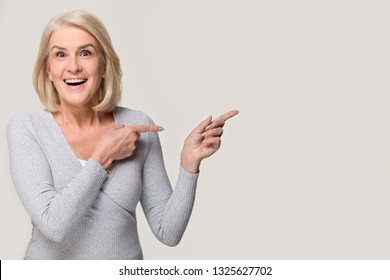 Amazed excited mature old woman pointing at copy space aside, happy surprised middle aged lady looking at camera advertising product service cheap price offer isolated on white grey studio background