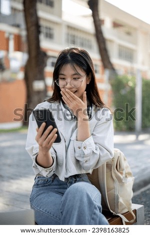 An amazed and excited young Asian woman is looking at her phone screen with a stunning face, covering her mouth, surprised with an unexpected news, sitting on a bench in a park. Stock photo © 