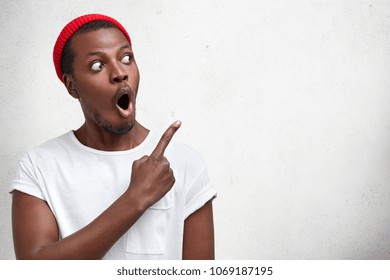 Amazed fashionable guy in red hat, keeps mouth widely opened, stares aside, indicates with fore finger at blank copy space, shows something strange and unexpected. Emotions and advertisment.