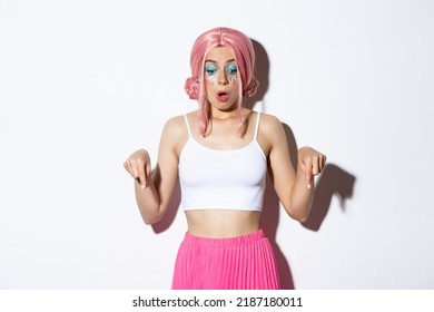 Amazed female model in pink wig and bright makeup, gasping in awe, pointing fingers down, looking at logo or banner, white background
