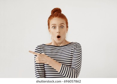 Amazed female model with red hair tied in knot, wearing stripe clothes, looking with bugged eyes and widely opened mouth, pointing with forefinger at copy space for your text or advertisment