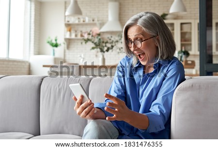 Amazed happy mature older 60s woman, excited customer holding smartphone using mobile app feeling great positive surprise reaction receiving gift reading sms on cell phone sitting on couch at home.