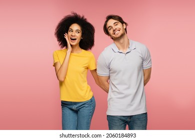 Amazed Hispanic Woman Touching Face Near Happy Man Hiding Something Behind Back Isolated On Pink