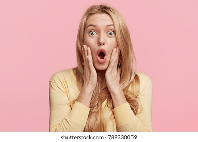 Amazed horrified female keeps mouth opened, hands on cheeks, being shocked to be explelled from university, stares at camera, isolated over pink background. Pretty woman expresses great surprisment