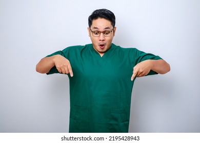 Amazed And Impressed Asian Male Nurse, Doctor In Scrubs Pointing Fingers Down And Looking Questioned, Interested In New Promo, White Background