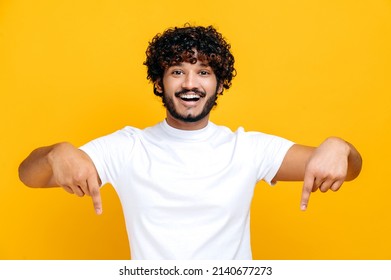Amazed indian or arabian cheerful young man in white basic t-shirt looks at the camera and points fingers down at space for your presentation, stands on isolated orange color background, smiling - Shutterstock ID 2140677273