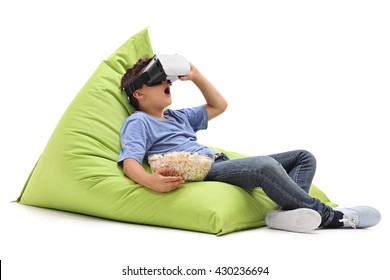 Amazed Little Boy Looking In VR Goggles And Eating Popcorn Seated On A Beanbag Isolated On White Background