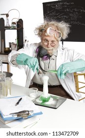 Amazed Mad Senior Scientist In Lab Reacts To Foaming Green Liquid Overflowing Beaker. Frizzy Grey Hair, Round Glasses, Lab Coat, Aqua Rubber Gloves, Blank Blackboard, Vertical, High Key, Copy Space.