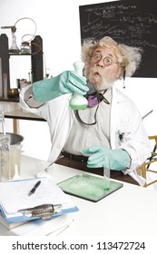 Amazed Mad Senior Scientist In Lab Reacts To Foaming Green Liquid Overflowing Beaker. Frizzy Grey Hair, Round Glasses, Lab Coat, Aqua Rubber Gloves, Blank Blackboard, Vertical, High Key, Copy Space.