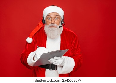 Amazed mature Santa Claus with real beard in red hat headset holding pen and clipboard, isolated on red background. Senior man planning his day, work and holiday celebration. - Powered by Shutterstock