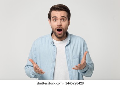 Amazed millennial man in blue shirt posing isolated on grey studio background, shocked guy open mouth makes big eyes cant believe in luck or surprised by offer raising spread palms feels impressed