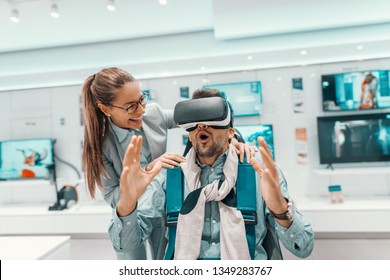 Amazed mixed race man sitting in chair and trying out vr technology. Woman standing next to him and holding his shoulders. Tech store interior. - Powered by Shutterstock