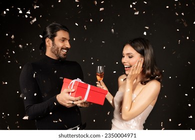 Amazed pretty young lady wearing elegant dress and nice makeup getting xmas present from her boyfriend, multiracial couple celebrating new year 2024 on black background, drinking champagne - Powered by Shutterstock