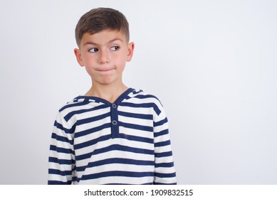 Amazed puzzled little cute Caucasian boy kid wearing stripped t-shirt against white wall, curves lips and has worried look, sees something awful in front.