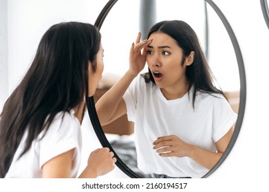 Amazed sad Chinese girl in a white t-shirt, standing in front of a mirror at home, looking at herself in the mirror in shock, saw a pimple or wrinkle, unpleasantly surprised, needs facial skin care
