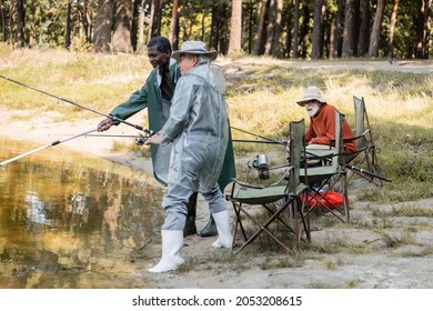 Amazed Senior Man Looking At Interracial Friends Fishing In Lake Water