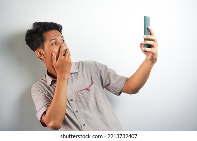 Amazed Shocked Asian Guy Holding Smartphone In His Hand, Looking At The Phone In Surprise, Stunned Facial Expression, Stands On Isolated White Background