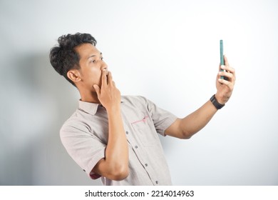 Amazed Shocked Asian Guy Holding Smartphone In His Hand, Looking At The Phone In Surprise, Stunned Facial Expression, Stands On Isolated White Background