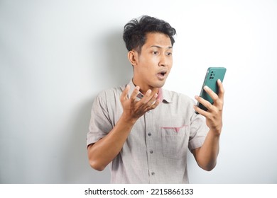 Amazed Shocked Asian Guy Holding Smartphone In His Hand, Looking At The Phone In Surprise, Stunned Facial Expression, Stands On Isolated White Background