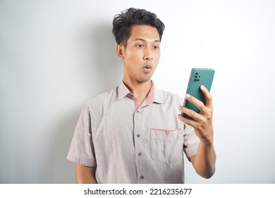 Amazed Shocked Asian Guy Holding Smartphone In His Hand, Looking At The Phone In Surprise, Stunned Facial Expression, Stands On Isolated White Background