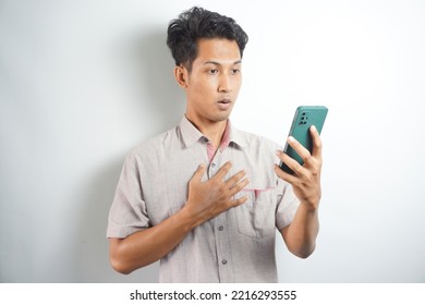 Amazed Shocked Asian Guy Holding Smartphone In His Hand, Looking At The Phone In Surprise, Stunned Facial Expression, Stands On Isolated White Background