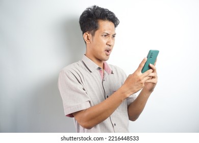 Amazed Shocked Asian Guy Holding Smartphone In His Hand, Looking At The Phone In Surprise, Stunned Facial Expression, Stands On Isolated White Background