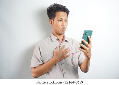 Amazed Shocked Asian Guy Holding Smartphone In His Hand, Looking At The Phone In Surprise, Stunned Facial Expression, Stands On Isolated White Background