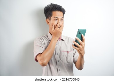 Amazed Shocked Asian Guy Holding Smartphone In His Hand, Looking At The Phone In Surprise, Stunned Facial Expression, Stands On Isolated White Background