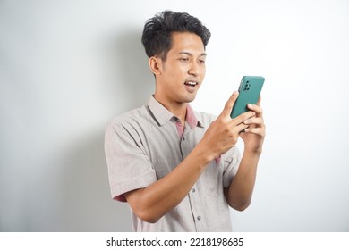 Amazed Shocked Asian Guy Holding Smartphone In His Hand, Looking At The Phone In Surprise, Stunned Facial Expression, Stands On Isolated White Background