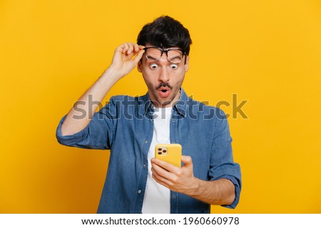 Amazed shocked caucasian guy holding smartphone in his hand, looking at the phone in surprise with his glasses raised, stunned facial expression, stands on isolated orange background