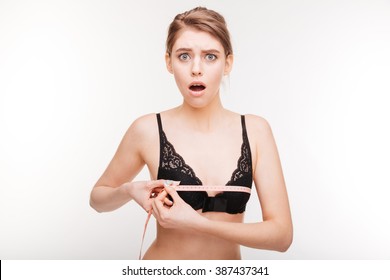 Amazed stunned young woman in black bra measuring her chest over white background