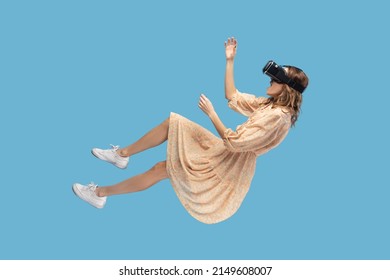 amazed surprised gamer girl hovering in air, levitating with virtual reality glasses on head, playing game through vr headset, floating in cyberspace. indoor studio shot isolated on blue background